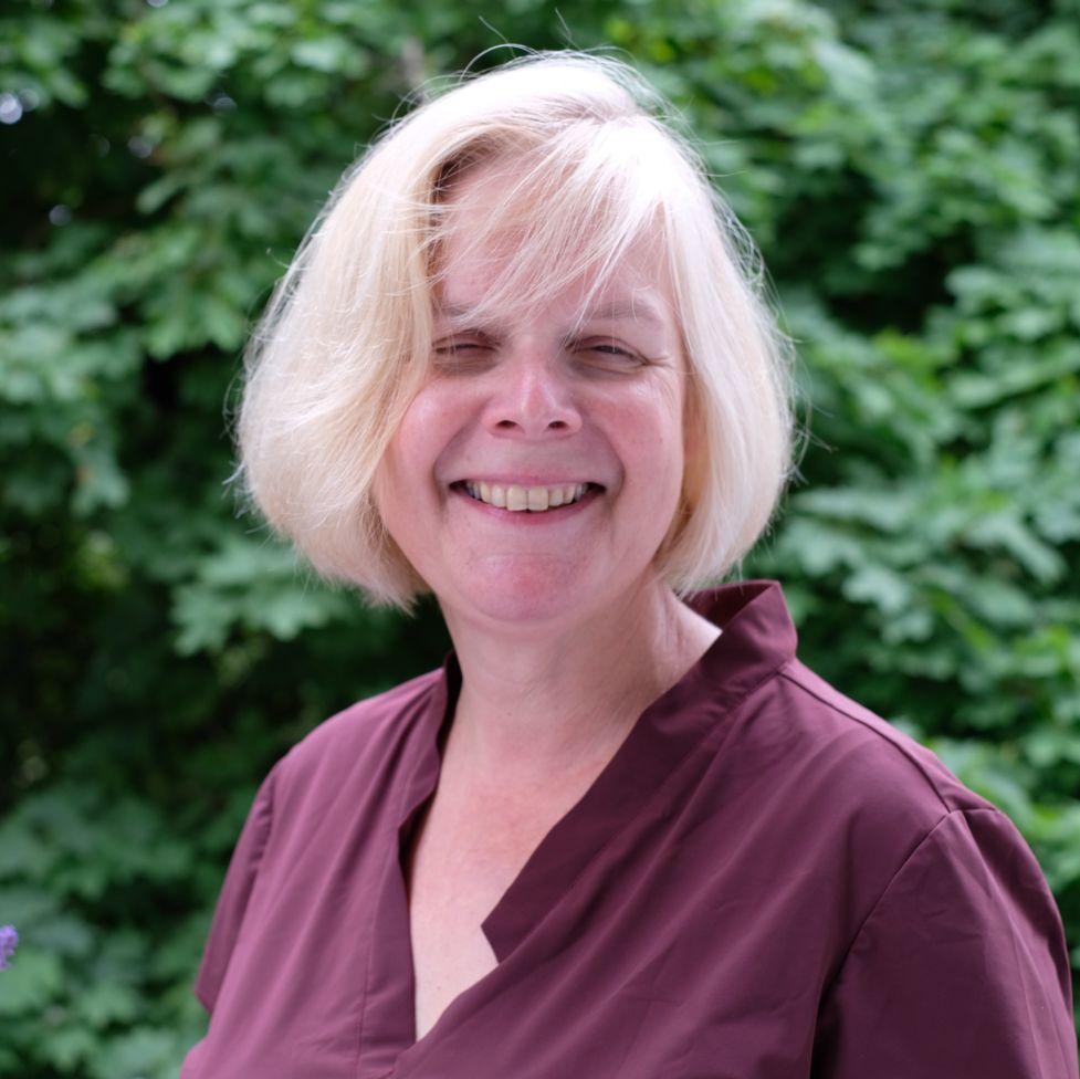 Dr.in Susanne Buchner-Sabathy, Expertin für digitale Barrierefreiheit, lächelt in die Kamera, hat blonde Haare, eine violette Bluse, im Hintergrund ein grüner Busch